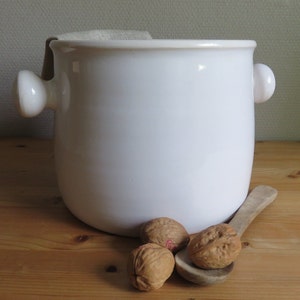 Puede incluir: Un tarro de cerámica blanca con dos asas y un forro de tela. El tarro está sentado sobre una superficie de madera con cuatro nueces a su lado.