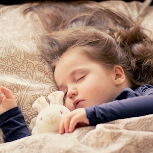 Può includere: Una bambina dorme profondamente nel letto, stringendo un coniglietto di peluche bianco. I suoi capelli sono sparsi sul cuscino e indossa una camicia blu scuro. Le lenzuola sono beige e marroni con un motivo.