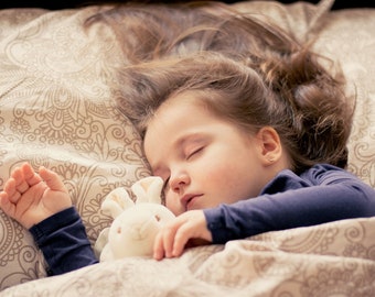 May include: A young girl sleeps soundly in bed, clutching a white plush bunny. Her hair is spread out on the pillow, and she is wearing a dark blue shirt. The bedding is a patterned beige and brown.