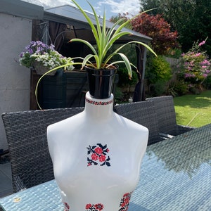 May include: A white ceramic bust with red and black floral embroidery, used as a planter for a green spider plant. The bust is sitting on a glass table with a wicker chair in the background.