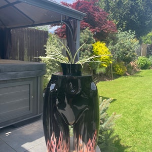 May include: A black planter shaped like a human torso with flames painted on the legs. A green plant is growing out of the top of the planter.