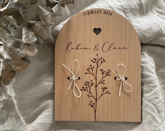 May include: Wooden arch-shaped sign with the text "13 Juillet 2024" and "Ruben & Clara" in cursive. The sign has two white strings tied around it, with a small heart cutout at the top.