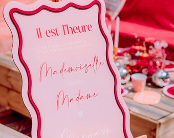 Peut inclure: Une pancarte rose avec une bordure blanche et un bord ondulé, sur un chevalet. La pancarte indique "Il est l'heure de boire du champagne et de monter sur les tables Mademoiselle devient Madame Margaux".