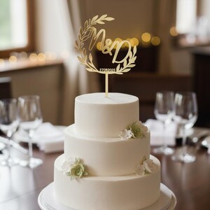 Può includere: Una torta nuziale bianca a tre piani con decorazioni floreali e un cake topper dorato. Il cake topper mostra le iniziali "M&D" all'interno di una corona d'alloro e la data "27.09.2025". La torta è su un vassoio bianco.