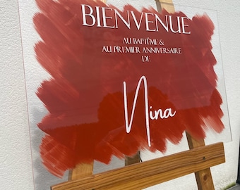 Peut inclure: Une pancarte en acrylique transparente avec un fond de couleur rouge en forme de coups de pinceau. La pancarte indique "Bienvenue au baptême & au premier anniversaire de Nina".