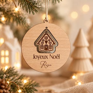 May include: A round, light-colored wooden ornament hanging from a string. It features a laser-cut gingerbread house design at the top, with the text "Joyeux Noël" and the name "Rose" engraved in dark brown lettering below the house.