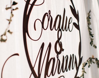 Peut inclure: Une pancarte de mariage en métal brun rustique avec un cadre circulaire et des accents de feuilles. La pancarte indique "Coralie & Marvin" en écriture cursive.