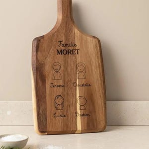 Könnte beinhalten: Holzschneidebrett mit Griff, graviert mit dem Familiennamen "Moret" und vier Strichmännchen mit Namen. Das Brett besteht aus hellem und dunklem braunem Holz und steht auf einer weißen Arbeitsplatte.