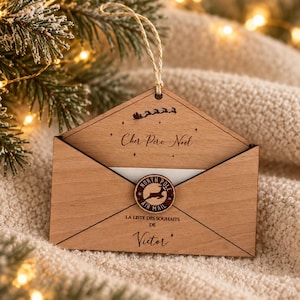 May include: A wooden envelope ornament featuring the text Cher Pere Noel, a silhouette of a sleigh, and a circular North Pole Air Mail seal. The envelope is engraved with the text LA LISTE DES SOUHAITS DE Victor and hangs from a gold string.