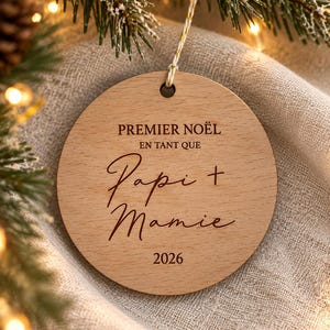 May include: A round, light-colored wooden ornament featuring the engraved text "PREMIER NOËL EN TANT QUE Papi + Mamie 2026". The ornament has a small hole at the top with a thin, twisted white and gold string attached for hanging purposes.