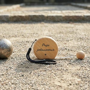 Peut inclure: Un mètre ruban en bois avec l'inscription "Papi pétancoeur" et un petit symbole de cœur. Une dragonne noire est attachée. Une boule métallique et une petite boule en bois sont également visibles sur une surface de gravier.