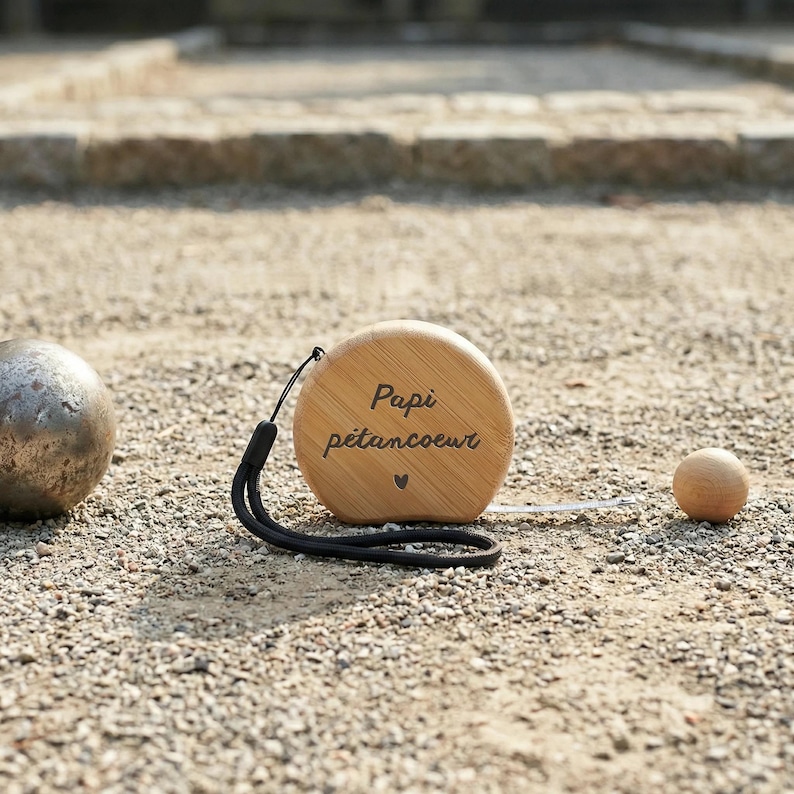 Peut inclure: Un m&egrave;tre ruban en bois avec l'inscription "Papi p&eacute;tancoeur" et un petit symbole de c&oelig;ur. Une dragonne noire est attach&eacute;e. Une boule m&eacute;tallique et une petite boule en bois sont &eacute;galement visibles sur une surface de gravier.