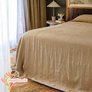 Peut inclure: Un couvre-lit en lin beige recouvre un lit, avec un oreiller assorti visible. Le lit est dans une pièce à la palette de couleurs neutres, avec une lampe et une table d'appoint en arrière-plan. Le couvre-lit a un aspect texturé.