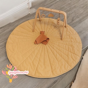 Puede incluir: Una alfombra de juego acolchada beige con un gimnasio de juego de madera en la parte superior. El gimnasio de juego tiene un anillo de madera colgante, un triángulo de madera y una cadena de cuentas de madera.