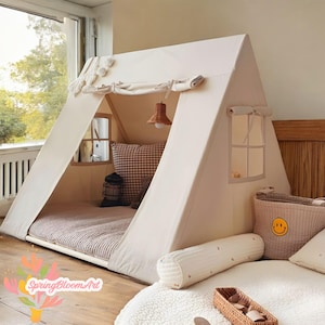 May include: A beige canvas play tent with a triangular roof and two window cutouts. Inside, a striped mattress and a checkered pillow are visible. A small woven basket and a smiley face basket are in front of the tent.