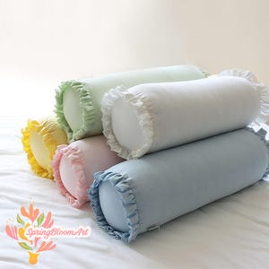 Puede incluir: Cinco almohadas decorativas de apoyo con bordes con volantes. Las almohadas están dispuestas en una pila, con la almohada más grande en la parte inferior y la más pequeña en la parte superior. Las almohadas son de color verde, amarillo, rosa y azul.