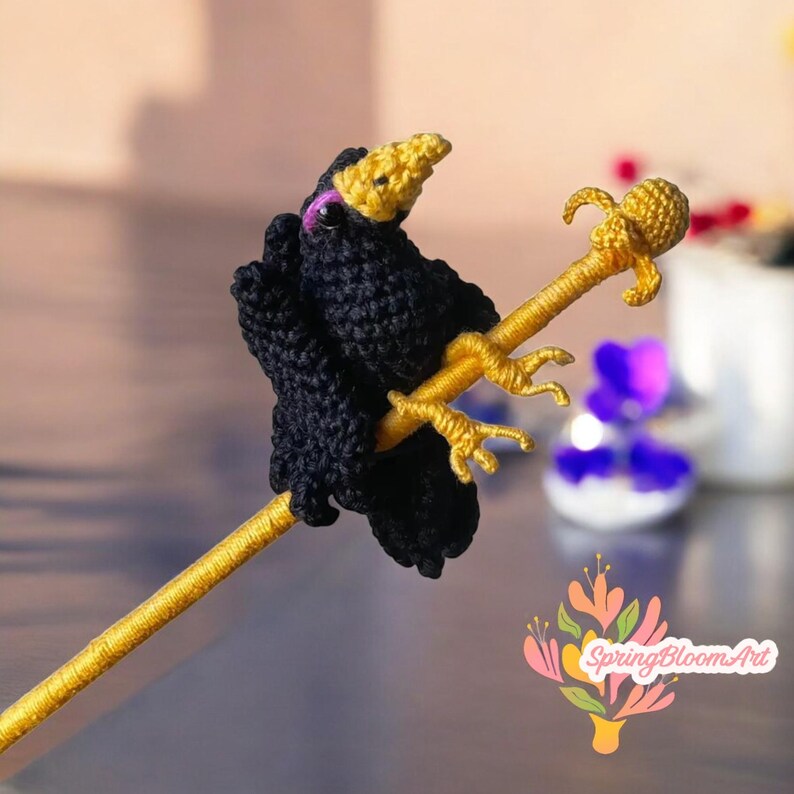 May include: A black crocheted bird perched on a gold stick with a gold flower on top. The bird has a pink eye and is holding onto the stick with its feet.