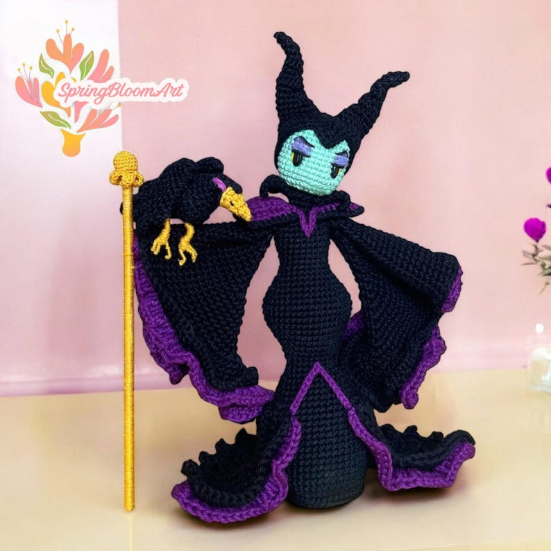May include: A crocheted Maleficent doll with purple and black clothing, holding a black raven in her right hand and a gold staff in her left hand. The doll has green eyes and a purple crown.