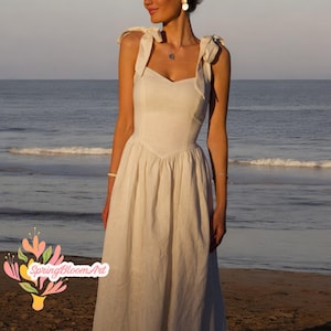 May include: A white linen dress with a sweetheart neckline and bow straps. The dress is photographed on a model standing on a beach.