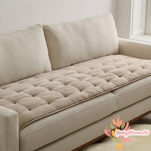 Può includere: Copricuscino per divano beige trapuntato. La fodera è rettangolare con una griglia di ciuffi quadrati. Il divano ha un tessuto beige chiaro e una base in legno. Anche i cuscini dello schienale sono beige.