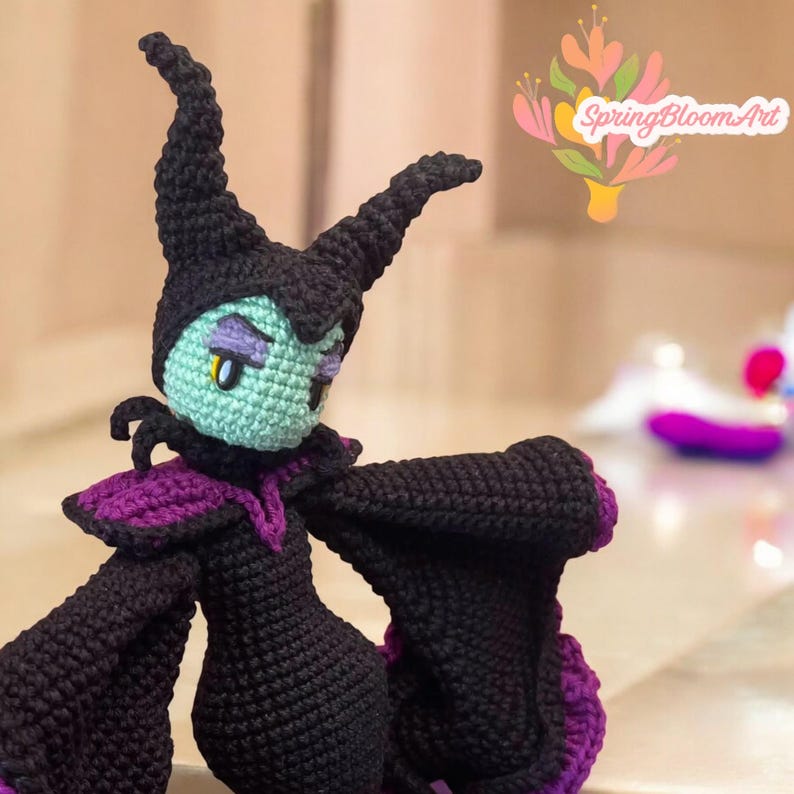 May include: A crocheted plush toy of Maleficent from Disney's Sleeping Beauty. The toy is green with purple accents and has black horns. The toy is wearing a purple cape.
