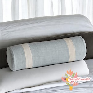 May include: A light blue bolster pillow with a beige stripe detail. The cylindrical pillow is made of a textured fabric and rests on top of other pillows and bedding. The background includes white curtains and other pillows.