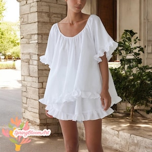 May include: A white linen two-piece set featuring a loose-fitting top with ruffled sleeves and hem, paired with matching shorts. The set has a scoop neckline and a flowing, lightweight design. The image is taken outdoors, with a brick wall and greenery in the background.