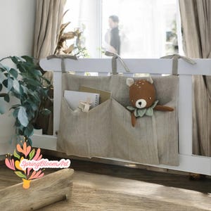 Può includere: Organizzatore appeso in lino beige con tre tasche. L'organizzatore è appeso a una ringhiera bianca. La tasca a sinistra contiene libri e una croce di legno. La tasca a destra contiene un giocattolo di peluche a forma di cervo.