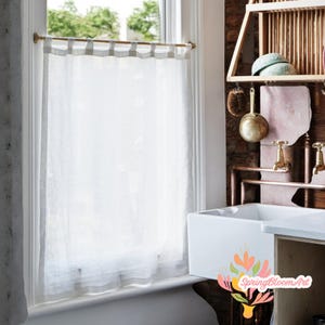 May include: White linen cafe curtain with a brass rod and rings. The curtain is hanging in a window with a view of trees and a building in the background. The curtain is partially open, revealing the view outside.