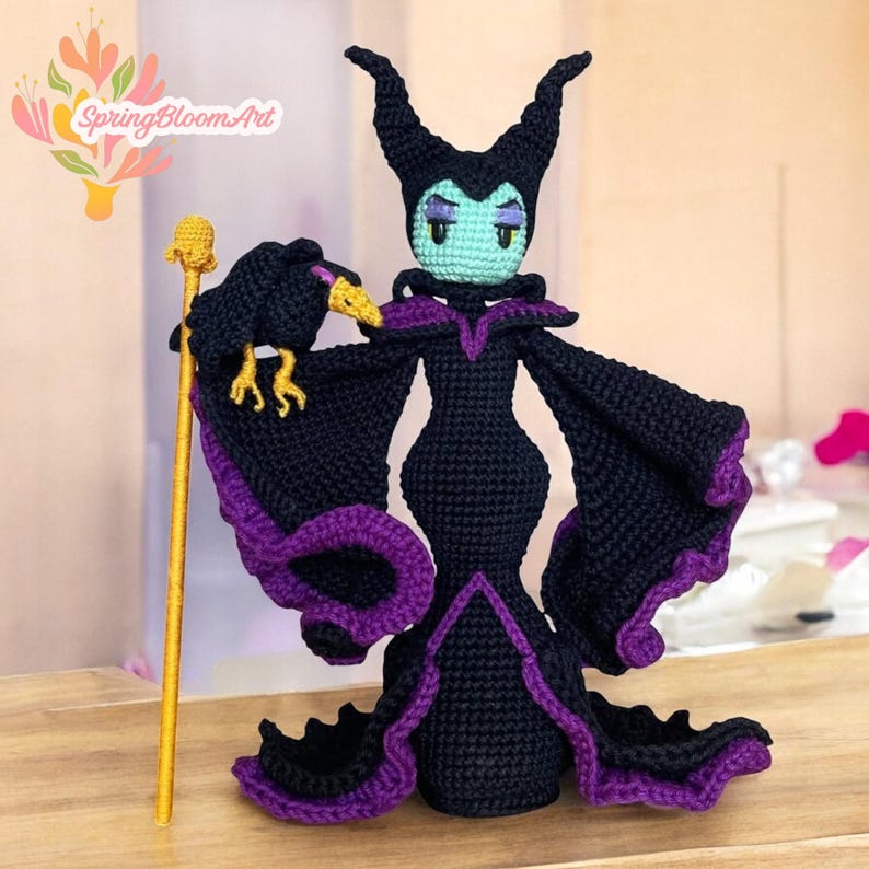 May include: A crocheted Maleficent doll with purple and black clothing, holding a black raven in her right hand and a gold staff in her left hand. The doll has green skin and purple eyeshadow.