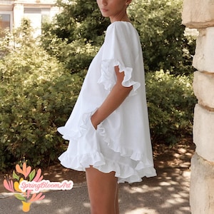 May include: White, ruffled mini dress with short sleeves and pockets. The dress features multiple layers of ruffles at the hem and sleeve openings. The dress is made of a lightweight fabric and is suitable for warm weather.