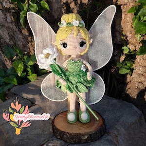 Könnte beinhalten: Eine gehäkelte Feenpuppe mit gelben Haaren, einem grünen Kleid und weißen Flügeln, die eine weiße Blume hält. Die Puppe steht auf einem Holzsockel.