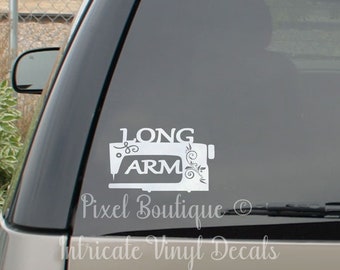 May include: White vinyl decal featuring a sewing machine design with the words "LONG ARM" and decorative floral elements. The decal is applied to a vehicle window. The text "Pixel Boutique" and "Intricate Vinyl Decals" are also visible.