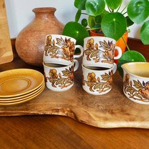 May include: Set of four floral patterned ceramic teacups and saucers with a matching creamer. The cups and saucers are stacked on a wooden chopping board.