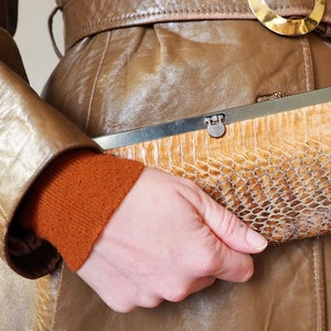 May include: A snakeskin-patterned clutch bag in tan and brown, with a silver clasp, is held against a brown leather coat. The coat has a gold buckle belt and an orange sweater.