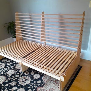 May include: A wooden slatted bed frame with a natural wood finish. The frame has a headboard and footboard with slatted panels. The frame is designed to be used with a futon mattress.