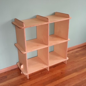 May include: A light-colored, wooden cube shelf unit with six open compartments. The shelf has a simple design with a rectangular shape and is made of unfinished wood. The unit is suitable for storage or display.
