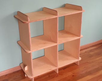 May include: A light-colored, wooden cube shelf unit with six open compartments. The shelf has a simple design with a rectangular shape and is made of unfinished wood. The unit is suitable for storage or display.