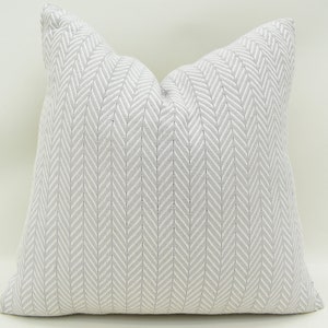 Puede incluir: Una almohada de tiro de color blanco y gris con un patrón de espiga. La almohada está hecha de una tela suave y texturizada.