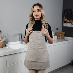 May include: A beige kitchen apron with a large pocket in the front. The apron has a textured woven fabric design.