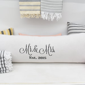 以下が含まれることがあります： 「Mr. & Mrs. Est. 2015」と黒色の刺繍が施された白い枕。枕は白い表面に置かれ、左側にグレーと白のストライプのタオルの積み重ね、右側にピンクと白のストライプのタオルの積み重ねがある。