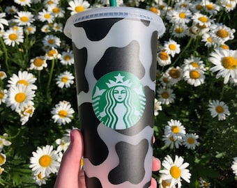 Può includere: Un bicchiere riutilizzabile con stampa di mucca bianco e nero, una cannuccia verde e il logo Starbucks. Il bicchiere è in un campo di margherite bianche.