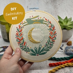 May include: A DIY embroidery kit featuring a circular design with a crescent moon, sunburst, and a rabbit. The embroidery uses green, yellow, orange, and red thread on a cream-colored fabric, held in a wooden hoop. The text "DIY Embroidery KIT" is in a yellow circle.