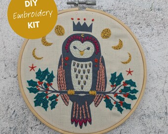 Könnte beinhalten: DIY-Stickset zum Sticken mit einer blauen und pinken Eule, die eine goldene Krone trägt und auf einem Zweig mit grünem Stechpalmenzweig und roten Beeren sitzt. Die Eule ist von goldenen Monden und Sternen umgeben.