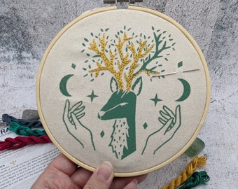 Könnte beinhalten: Stickerei auf einem Reifen mit einem grünen Hirsch mit einer gelben und grünen Blumenkrone. Der Hirsch ist von zwei Händen und Sternen auf einem cremefarbenen Hintergrund umgeben.