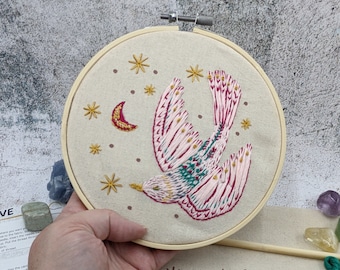 Könnte beinhalten: Ein gestickter Vogel in Rosa und Grün, der mit einer Sichelmond und Sternen auf einer weißen Leinwand in einem hölzernen Stickrahmen fliegt.