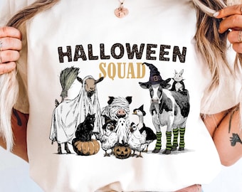 May include: Cream-colored t-shirt featuring a Halloween Squad design. The graphic includes a goat in a sheet, a black cat, a pig wrapped in bandages, a cow in a witch hat, and other spooky characters. The text "HALLOWEEN SQUAD" is displayed above the characters.
