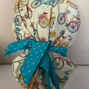 May include: A white scrub hat with a turquoise polka dot tie. The hat features a repeating pattern of vintage bicycles with flowers.