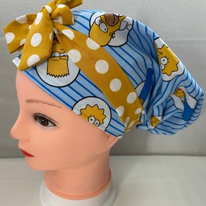 May include: A blue and yellow surgical scrub hat with a yellow bow and a pattern of cartoon characters from the Simpsons. The hat is on a white mannequin head.