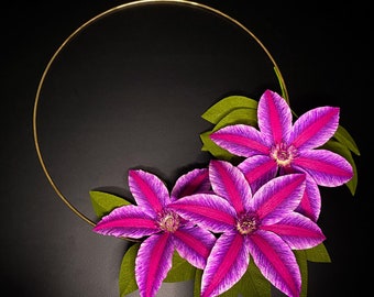 May include: A gold metal hoop with three purple and white paper flowers with green leaves. The flowers have a striped pattern and are arranged in a cluster.